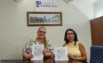 Two women sit at a table holding printed documents, smiling at the camera inside an office with a Puerto Paraiso sign on the wall.