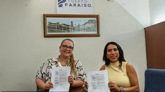 Two women sit at a table holding printed documents, smiling at the camera inside an office with a Puerto Paraiso sign on the wall.