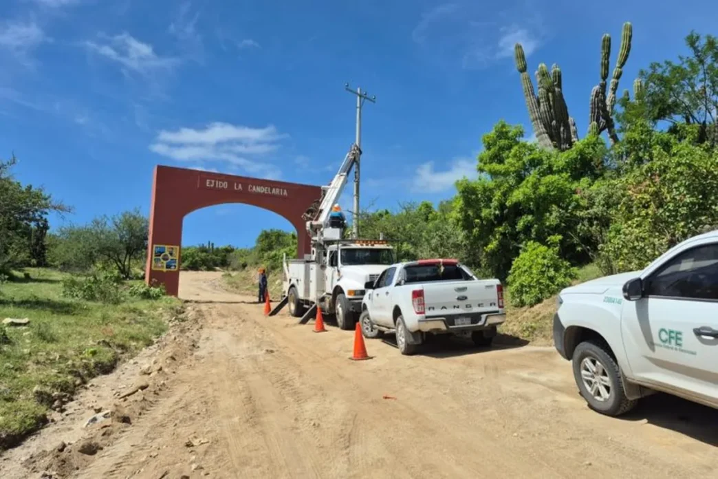 .5M Project Brings Power to La Candelaria, Puerto Chale - Gringo Gazette Dirt road work scene with a bucket truck, orange cones, and trucks near a red arch that says 'Ejido La Candelaria' in a rural area.