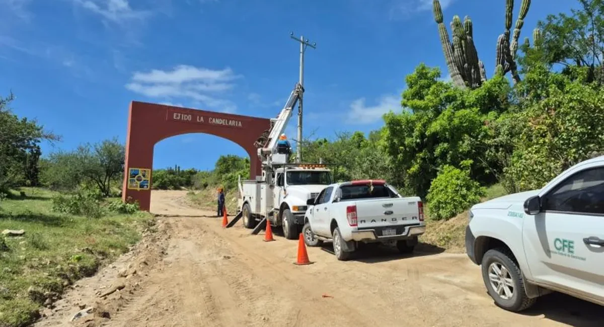 Dirt road work scene with a bucket truck, orange cones, and trucks near a red arch that says 'Ejido La Candelaria' in a rural area.