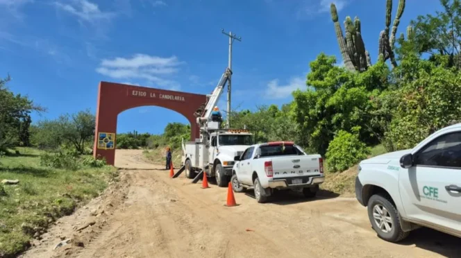 Dirt road work scene with a bucket truck, orange cones, and trucks near a red arch that says 'Ejido La Candelaria' in a rural area.