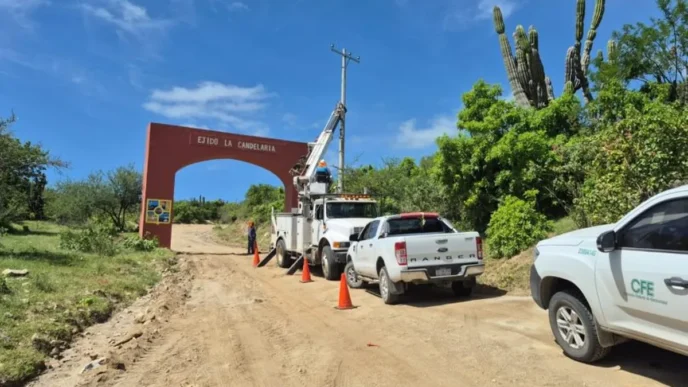Dirt road work scene with a bucket truck, orange cones, and trucks near a red arch that says 'Ejido La Candelaria' in a rural area.